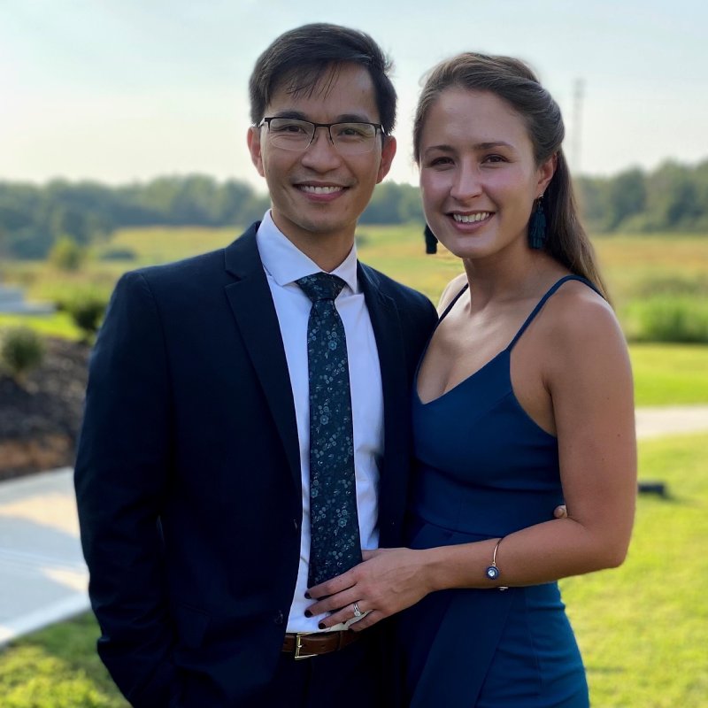 A smiling couple posing outdoors: man in a dark suit and patterned tie, woman in a blue dress with teal earrings, standing close together and holding hands in a sunlit park.