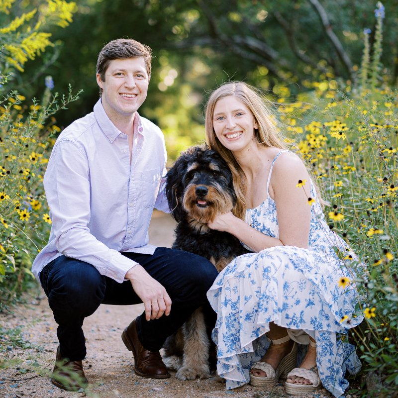Smiling couple kneels in a sunny garden with a fluffy dog between them, yellow wildflowers surrounding them