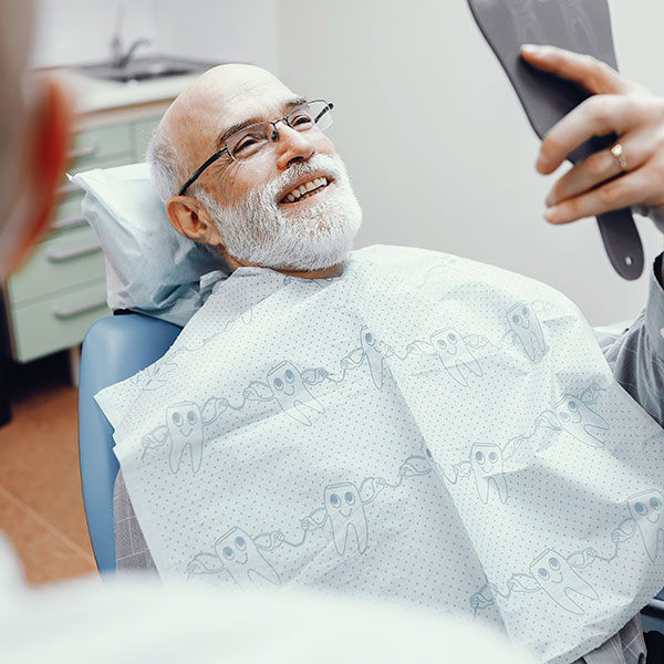 WhattoExpect Older man with glasses reclines in a dental chair, smiling as a dentist uses a mirror to inspect his teeth.